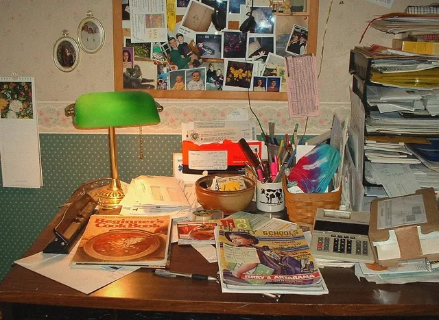 Un bureau encombré : lampe verte, magazines, pots de crayons, photos punaisées au mur (photo provisoire).